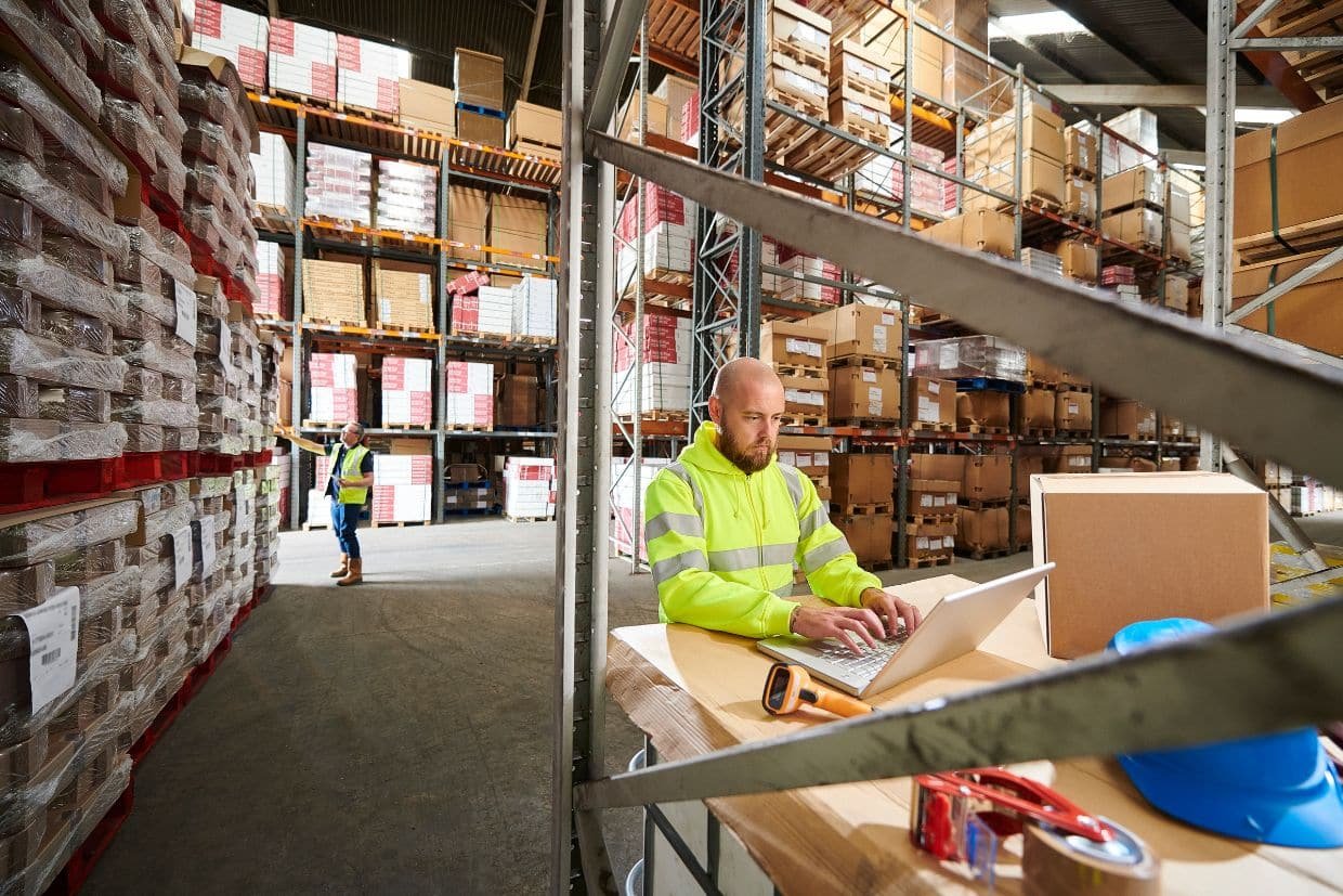 Warehouse worker in a high-visibility jacket using a laptop at a workstation inside a large warehouse filled with stacked pallets and shelves of packaged goods. Another worker in the background is inspecting inventory
